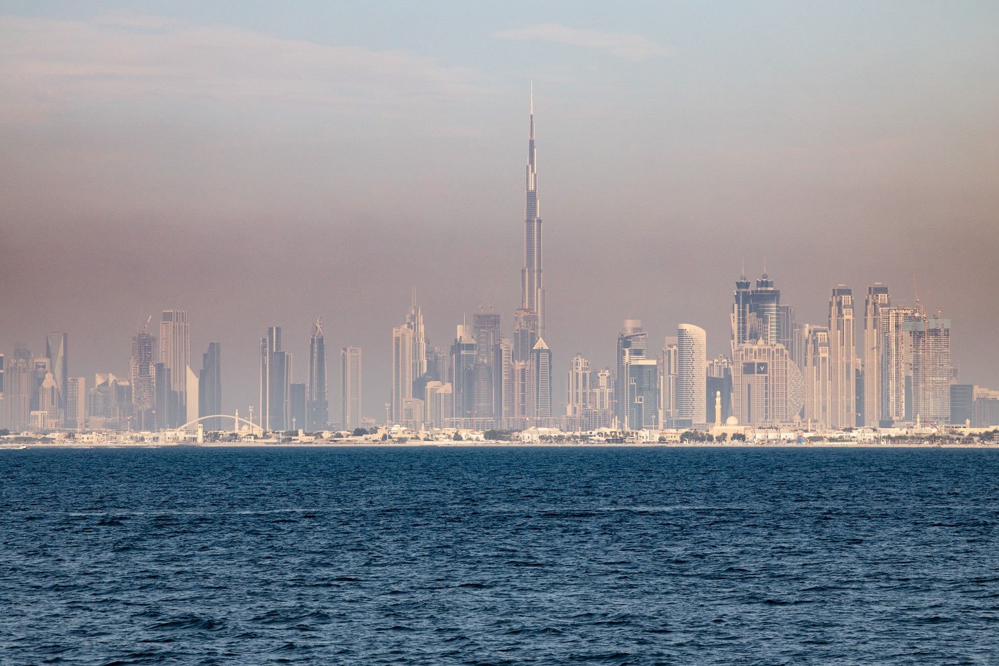 View across the sea toward the Dubai skyline with the Burj Khalifa towering above other skyscrapers under a hazy sky.
