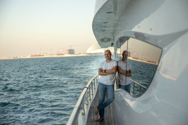 A man in a white shirt standing on the railing of a large white yacht, with the sea and coastal skyline visible in the background.