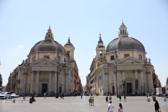 Two nearly identical domed Baroque churches flank a street entrance in a Rome piazza, with people walking across the cobblestone square.