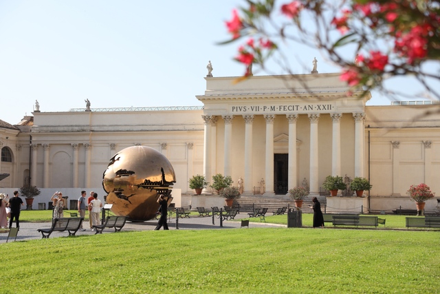 A golden cracked-sphere sculpture stands on a lawn in front of a neoclassical building with Corinthian columns at the Vatican Museums, with visitors nearby and red flowers framing the foreground.