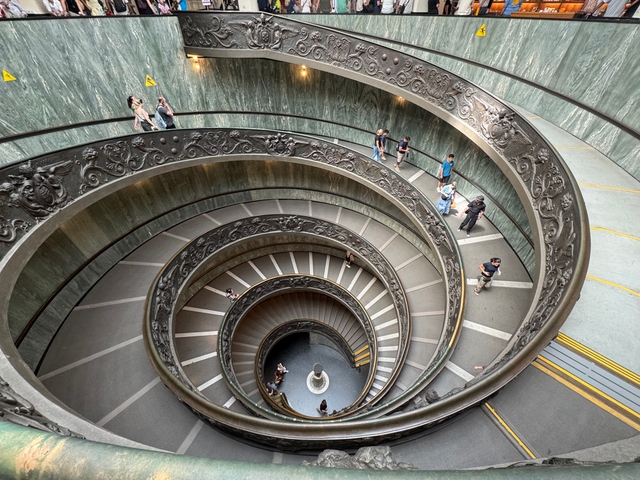 A double-helix spiral staircase with ornate bronze railings seen from above, with visitors walking along its curving steps.
