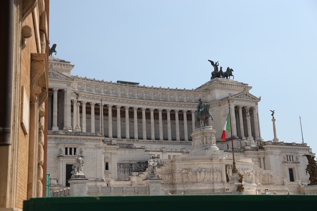 View of the white marble Altare della Patria monument in Rome with its columns, bronze quadriga statues, and an Italian flag against a clear blue sky.