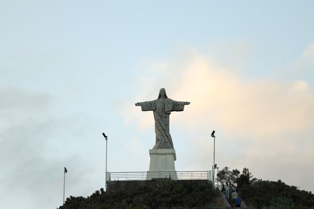 A tall stone statue of Christ with arms outstretched stands atop a hill in Madeira, with visitors climbing steps toward it under a pale evening sky.